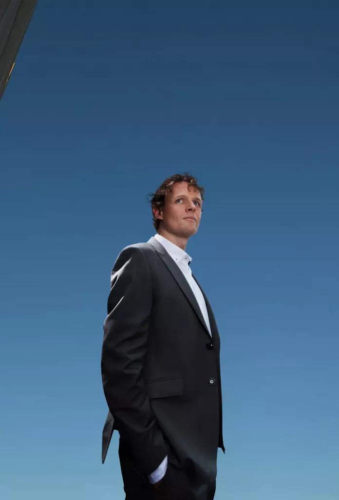 Man in een donker pak staat met een hand in zijn zak tegen een diepblauwe lucht