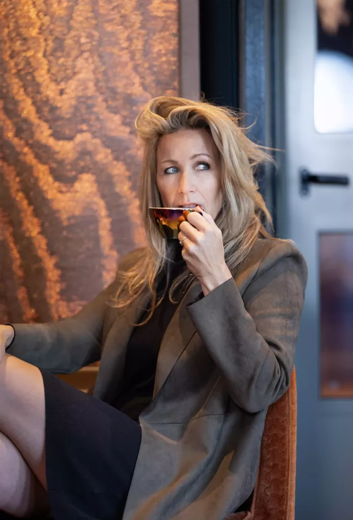 Vrouw met blond haar in een bruin colbert zit bij een raam met een glas aan haar lippen