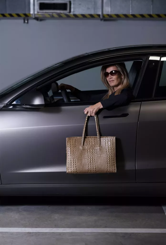 Vrouw met zonnebril leunt uit het raam van een grijze auto en houdt een bruine tas naar buiten