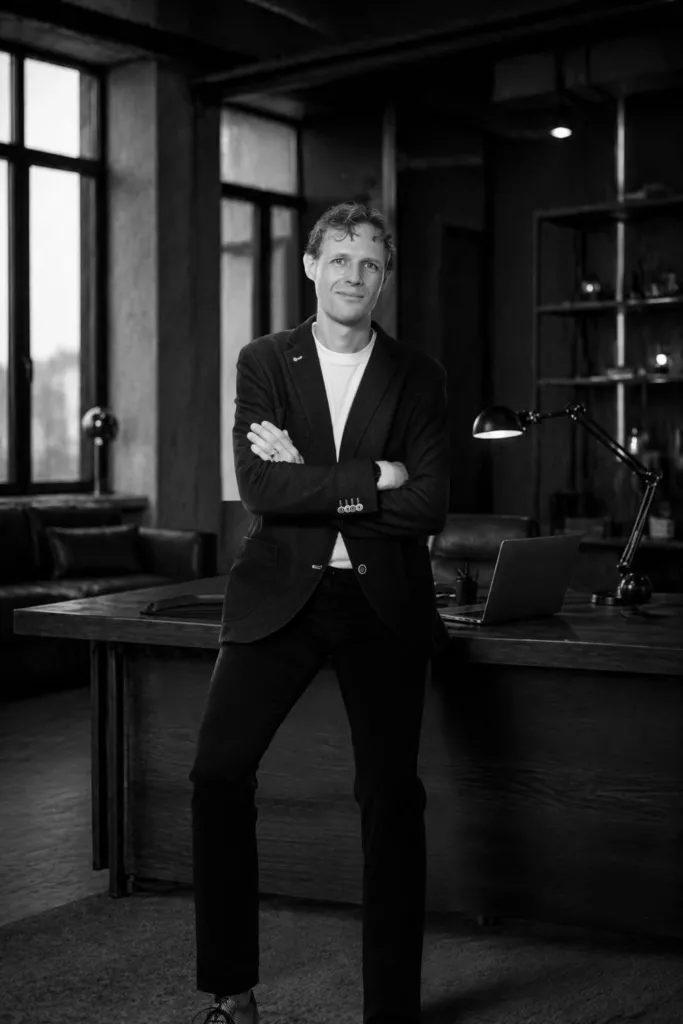 Zwart-witportret van Floris Koolen met gekruiste armen, leunend tegen een bureau in een donker kantoor