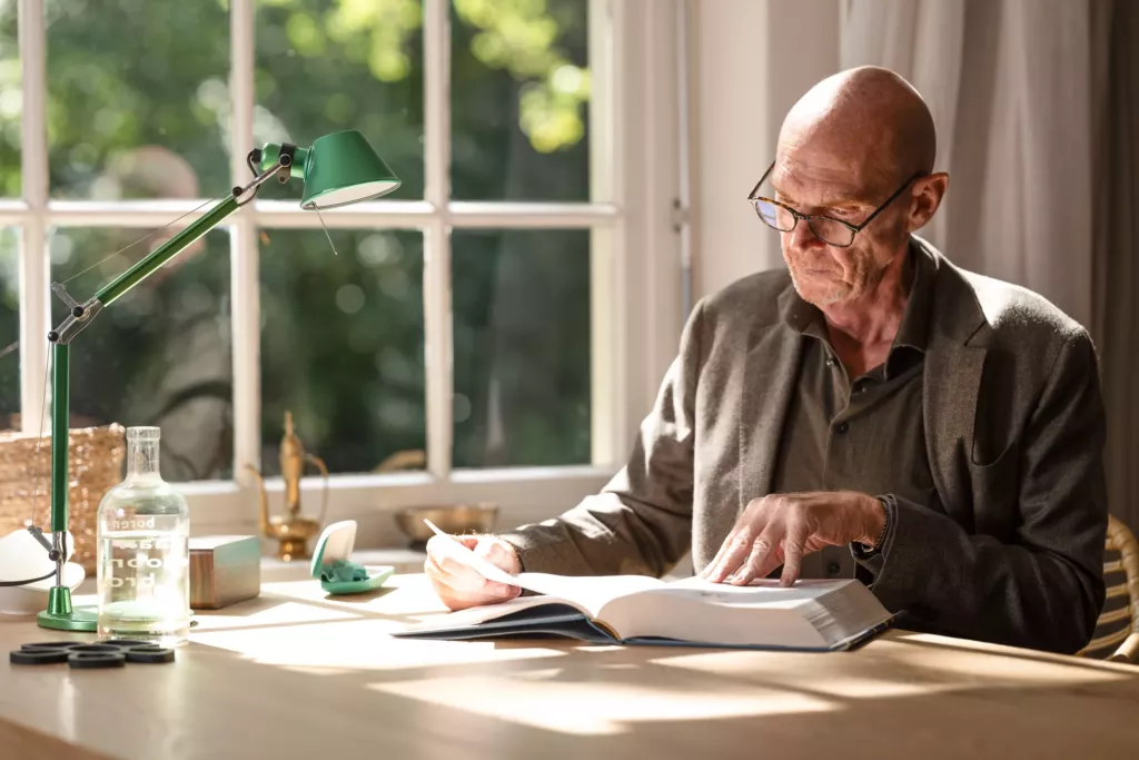 Peter, een man met grijs haar, zit aan een tafel bij een raam en kijkt in een open boek.