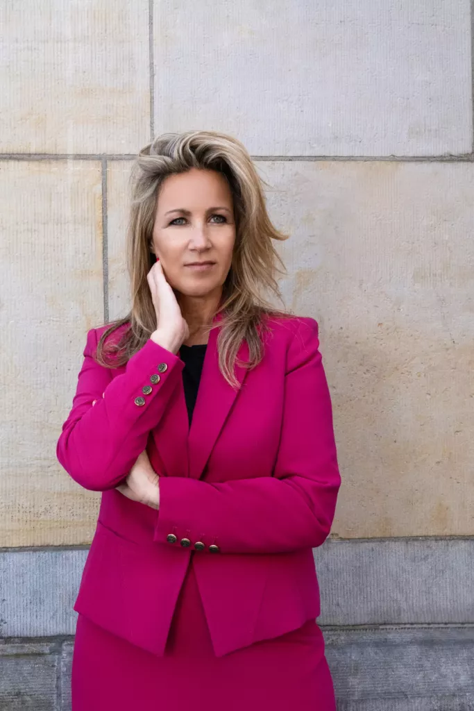 Meriam staat in een felroze blazer tegen een stenen muur.